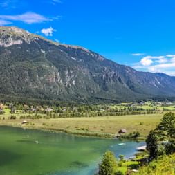 Pressegger See mit grünem Wasser, Wiesen und bewaldeten Bergen unter blauem Himmel. Kleines Dorf und Stege am Ufer sichtbar.
