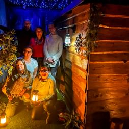 Six team members posing at a wooden Christmas market hut decorated with lights. Blue starry ceiling and festive decorations visible.