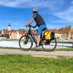 Cyclist with yellow panniers riding along the Lech river in Landsberg am Lech, with historic buildings and church tower in background.