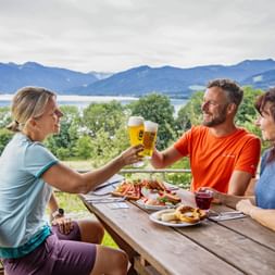 Three cyclists toasting with beer at a wooden table with food, overlooking Tegernsee lake and mountains. Bike helmets visible on the table.