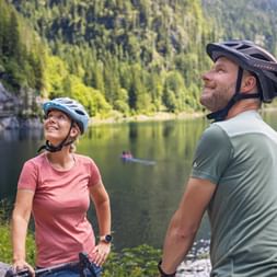 Two cyclists with helmets pausing by Gosausee lake in Salzkammergut, surrounded by forested mountains and calm water reflecting the scenery.