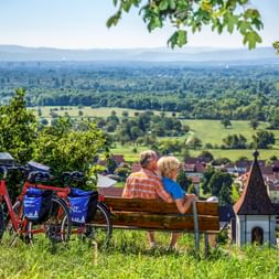 Two cyclists sitting on wooden bench overlooking Istein village in Southern Black Forest. Two bikes with blue panniers parked nearby, green valley below.