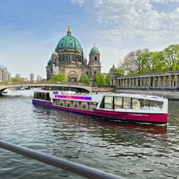 Purple and white tour boat on the Spree River with Berlin Cathedral's green dome visible in the background under a partly cloudy sky.
