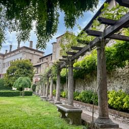 Garden of Castello del Buonconsiglio in Trento with stone pergola, manicured lawn, and historic castle building under blue sky.