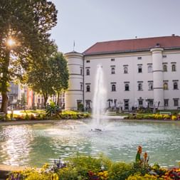 Brunnen vor Schloss Porcia in Spittal mit bunten Blumenbeeten. Das weiße Renaissance-Schloss hat ein rotes Dach und einen Turm.