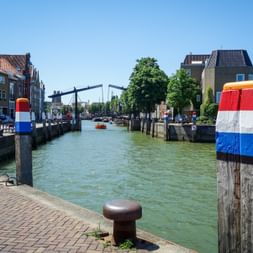 Canal in Dordrecht with mooring posts painted in Dutch flag colors, historic buildings, and a drawbridge under blue sky.