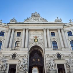 White baroque facade of the Hofburg Palace in Vienna with ornate columns, sculptures, and a grand arched entrance under a clear blue sky.