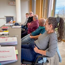 Group of six people seated in a bright meeting room with large windows, attending a training session. Documents labeled 'Eurofun Akademie' are visible on the table.
