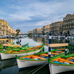 Traditionelle weiße Ruderboote mit grünem und orangem Rand im Kanal von Sète. Historische Gebäude säumen das Ufer unter blauem Himmel.