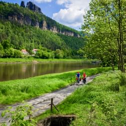 Two cyclists riding along a paved path beside the Elbe River with green meadows, forested cliffs, and houses in the background under blue sky.