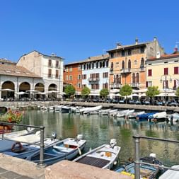 Small harbor in Desenzano at Lake Garda with moored boats, colorful buildings, and white arcaded structure under clear blue sky.