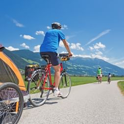 Cyclist with child trailer on paved path through Austrian countryside, mountains in background. Other cyclists visible ahead on sunny day.