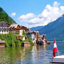 View of Hallstatt village from a boat with Austrian flag. Traditional Alpine houses line the lakefront with mountains in background.