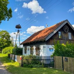 Traditionelles masurisches Haus mit Holzfassade und orangefarbenem Ziegeldach in Wojnowo. Ein Storchennest sitzt auf einem Pfahl neben dem Haus, davor ein Holzzaun.