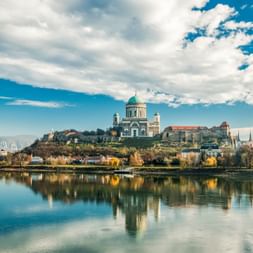Esztergom Basilica with green dome on hilltop overlooking the Danube River. Historic buildings and castle ruins visible, with reflections in calm water.