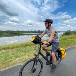 Cyclist with yellow panniers riding along paved path beside Loing canal. Green grass and trees line the waterway under a blue sky with clouds.