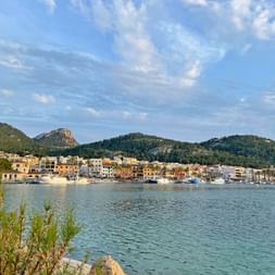 Hafen Port d'Andratx auf Mallorca mit bunter Häuserfront am Wasser, Booten im ruhigen Wasser und grünen Hügeln unter blauem Himmel.