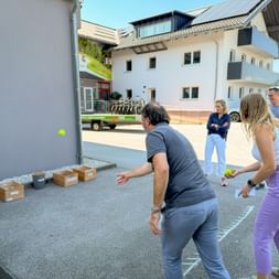Four people playing an outdoor game with cardboard boxes and tennis balls on pavement. Modern white building with solar panels in background.