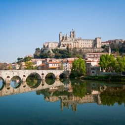 Steinbrücke mit Bögen spiegelt sich im ruhigen Flusswasser, mit Kathedrale Saint-Nazaire und historischen Gebäuden auf Hügel in Béziers.
