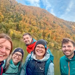 Five smiling people in outdoor jackets taking a selfie by the Danube river with autumn-colored hills in the background during a company trip.