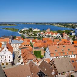 Aerial view of Stralsund's old town with orange-tiled roofs and half-timbered houses overlooking a blue harbor with sailboats and green shores.