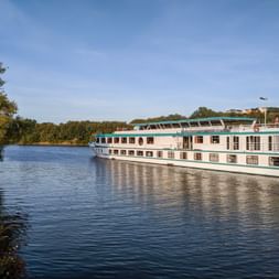 MS Switzerland river cruise ship with white hull and turquoise trim sailing on calm river, surrounded by green trees under blue sky.