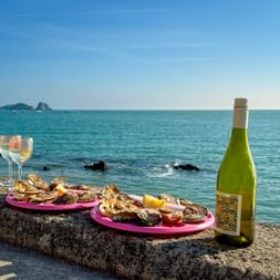 Fresh oysters on pink plates with white wine glasses and bottle on a stone wall overlooking the turquoise sea in Cancale, Brittany.