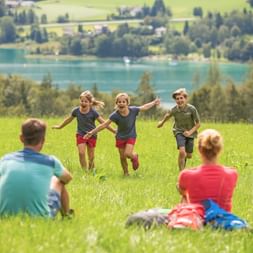 Three children running through green meadow toward parents during picnic. Turquoise alpine lake and rolling hills visible in background.