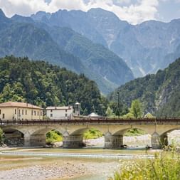 Historische Steinbrücke mit mehreren Bögen über einen Fluss in Resiutta, mit traditionellen Gebäuden und dramatischen Berggipfeln im Hintergrund.