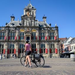 Radfahrerin mit Fahrrad vor dem historischen Rathaus von Delft, einem Renaissance-Gebäude mit roten Fensterläden unter blauem Himmel.