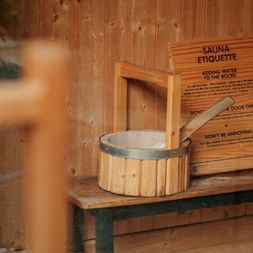 Wooden sauna interior with etiquette sign on wall, wooden bucket with ladle on bench, and blurred person in foreground.