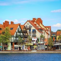 Bunte Ufergebäude mit orangen Ziegeldächern in Mikołajki, Masuren. Fachwerk- und traditionelle Architektur säumt den See unter blauem Himmel.