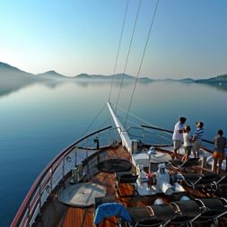 Bow deck of MS Dalmatino with passengers enjoying calm morning waters. Misty Croatian coastline and mountains visible in the background.