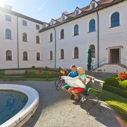 Zwei Radfahrer auf einer Bank im Innenhof des Klosters Seeon mit Brunnen, gepflegten Gärten und weißem Klostergebäude mit Bogenfenstern.