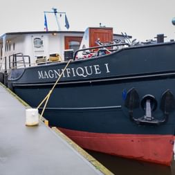 Flusskreuzfahrtschiff MS Magnifique I mit dunkelblauem Rumpf und rotem Boden am Kai festgemacht. Fahrräder auf Deck sichtbar, weißer Aufbau mit Fenstern.