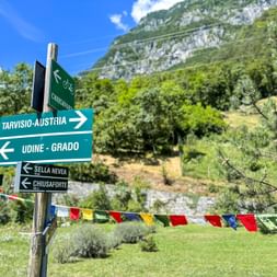 Wegweiser für Radfahrer nach Tarvisio-Österreich und Udine-Grado im Kanaltal, mit Bergen und bunten Wimpeln im Hintergrund.