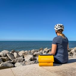 Radfahrerin mit weißem Helm sitzt auf der Promenade in Lido di Venezia und blickt aufs Meer. Gelbe Eurobike-Box neben ihr an felsiger Küste.