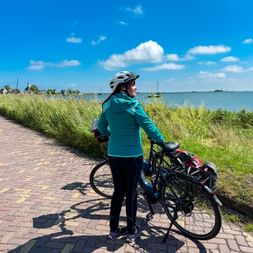 Frau mit Fahrrad auf gepflastertem Weg am IJsselmeer. Sie trägt türkisfarbene Jacke und Helm und blickt auf Wasser mit Booten und blauem Himmel.