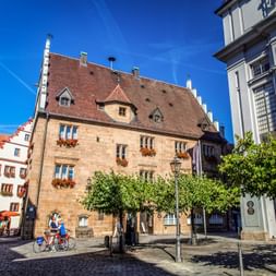 Two cyclists with bicycles on a cobblestone square in Ansbach, surrounded by historic buildings with red-tiled roofs and green trees.