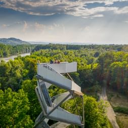Moderner Aussichtsturm mit Zickzack-Plattformen über dichtem Wald. Die Mur schlängelt sich durch grüne Landschaft unter blauem Himmel.