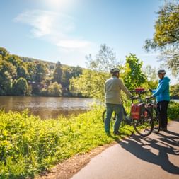 Two cyclists with loaded touring bikes pause on a riverside path along the Werra-Fulda route, surrounded by green vegetation and forested hills.