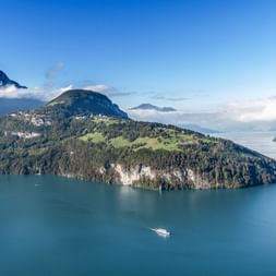 Luftaufnahme des Vierwaldstättersees mit einer Halbinsel mit grünen Wiesen und Wäldern. Berge erheben sich im Hintergrund, ein Passagierboot fährt.