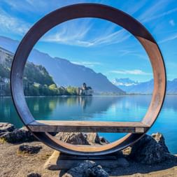 Kreisförmige Metallskulptur rahmt Blick auf Schloss Chillon am Genfersee. Berge und blauer Himmel im Hintergrund. Montreux Uferpromenade.