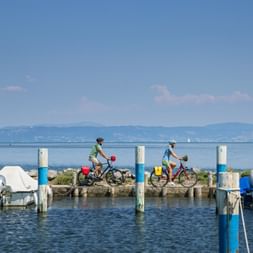 Two cyclists riding along a wooden pier in Grado harbor with boats moored on both sides, calm blue water, and mountains in the distance.