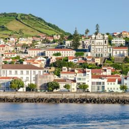 Küstenansicht der Altstadt Horta auf Faial mit bunten Gebäuden, Kirchen und grünem Hügel. Eine Steinmauer grenzt an das blaue Wasser.