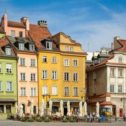 Reihe bunter historischer Gebäude am Warschauer Marktplatz mit roten Ziegeldächern. Außengastronomie mit weißen Sonnenschirmen davor.