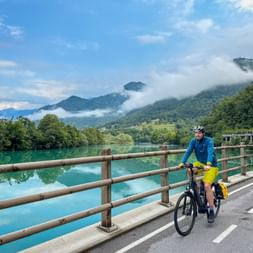 Cyclist in blue jacket and yellow shorts riding on bridge over turquoise Soča River with misty mountains in background, Slovenia.
