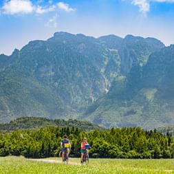 Two cyclists riding on a path through green meadow near Salzburg-Wals with Untersberg mountain in background under blue sky.