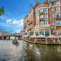 Red brick buildings with ornate facades line the Amstel River in Amsterdam. A boat travels on the water, and outdoor terraces are visible along the canal.