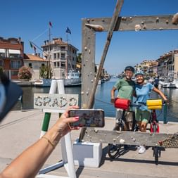 Two cyclists posing with bikes at Grado harbor photo frame while another person takes their picture with a smartphone.